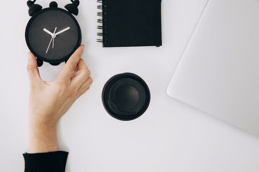 close up person s hand holding black alarm clock spiral notepad laptop takeaway coffee cup white desk